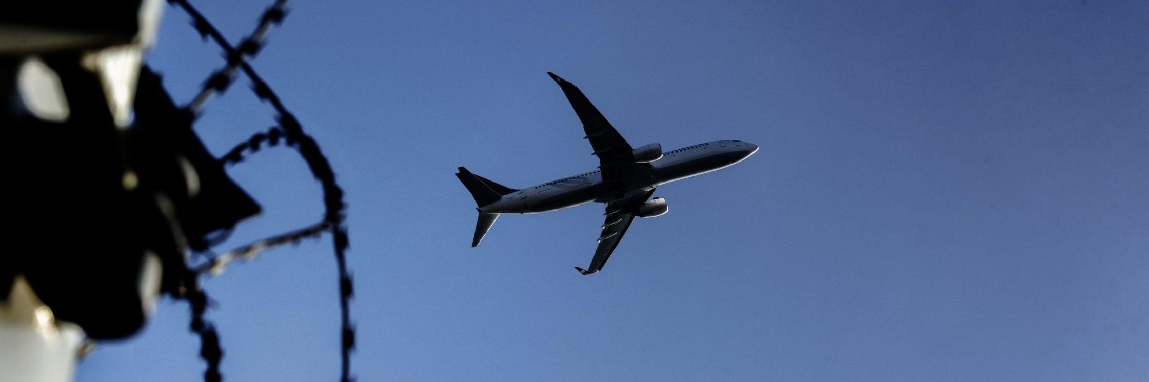 Ein Flugzeug fliegt über Stacheldraht vor blauem Himmel