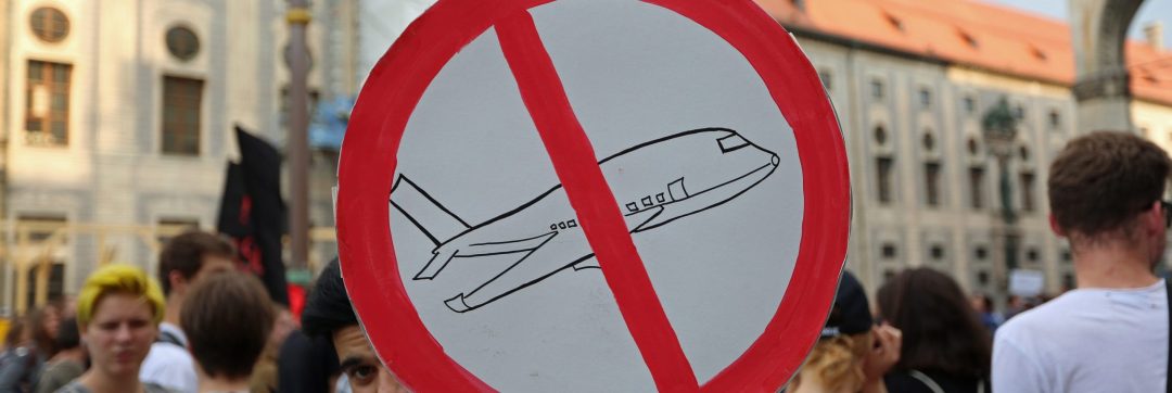 Pappschild mit einem durchgestrichenen Flugzeug im Verbotszeichen. Im Hintergrund sind Menschen, die protestieren.